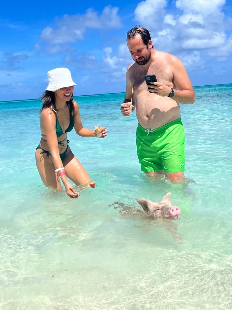 A happy couple on the beach in the Bahamas, surrounded by crystal-clear turquoise waters. They are feeding friendly swimming pigs, adding to their unforgettable trip.